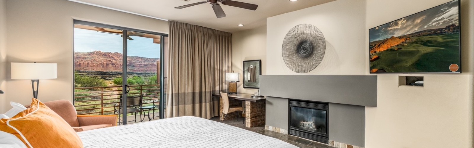 A cozy bedroom with a large bed, TV, fireplace, desk, and a scenic view outside through sliding glass doors, complete with a ceiling fan.