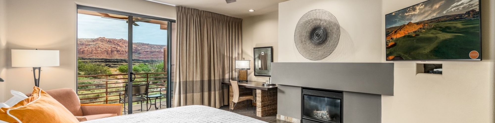 A cozy bedroom with a large bed, TV, fireplace, desk, and a scenic view outside through sliding glass doors, complete with a ceiling fan.