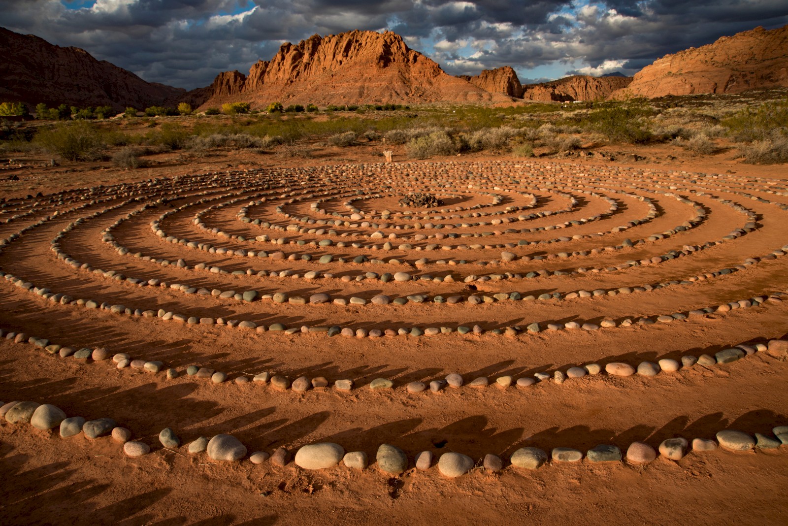 A circular labyrinth made of stones in the desert with a mountainous backdrop and a partially cloudy sky.