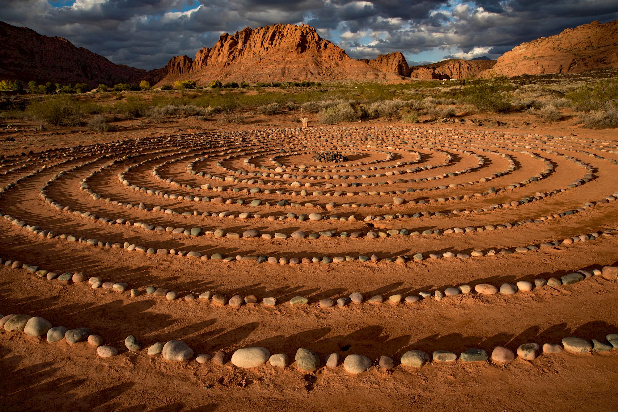 A circular labyrinth made of stones in the desert with a mountainous backdrop and a partially cloudy sky.