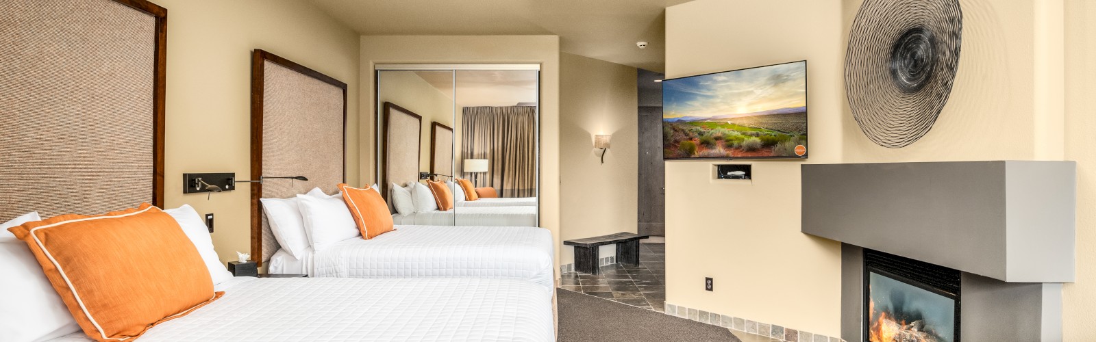 A hotel room features two beds with orange pillows, a ceiling fan, a fireplace, a TV, and a large mirror. The floor has a mix of carpet and tiles.