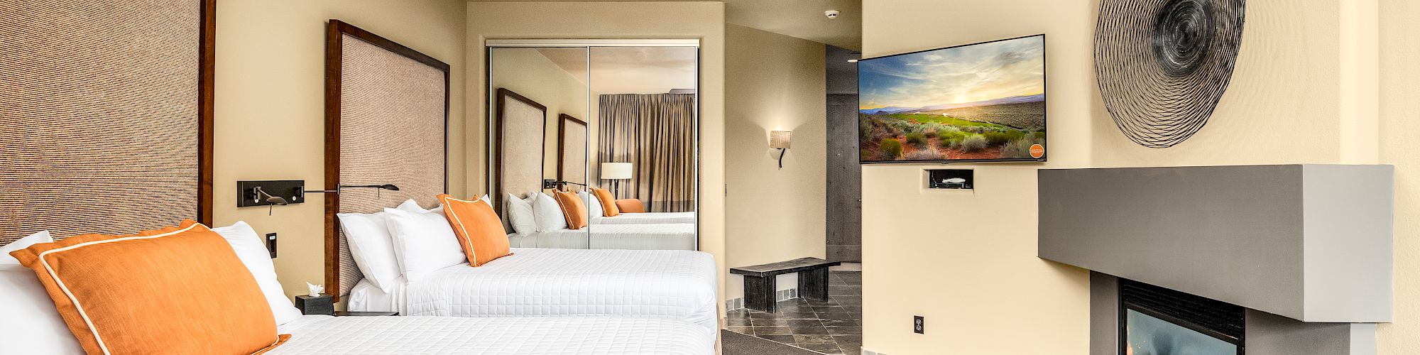 A hotel room features two beds with orange pillows, a ceiling fan, a fireplace, a TV, and a large mirror. The floor has a mix of carpet and tiles.