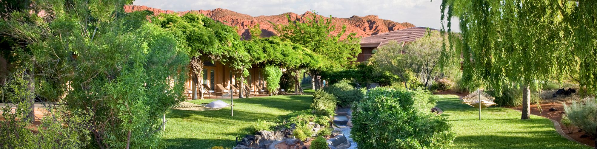 A beautiful garden with a stream flowing through, surrounded by lush green trees, plants, and manicured lawns, with red rock hills in the background.