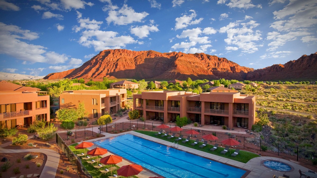 A scenic resort with an outdoor pool and sun loungers, surrounded by desert landscape and red rock mountains under a blue, partly cloudy sky.