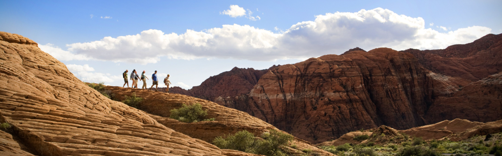 A group of people hiking on rocky terrain under a blue sky with scattered clouds, surrounded by a scenic mountain landscape and greenery.