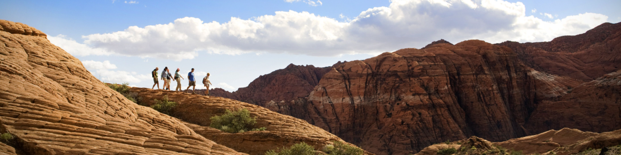 A group of people hiking on rocky terrain under a blue sky with scattered clouds, surrounded by a scenic mountain landscape and greenery.