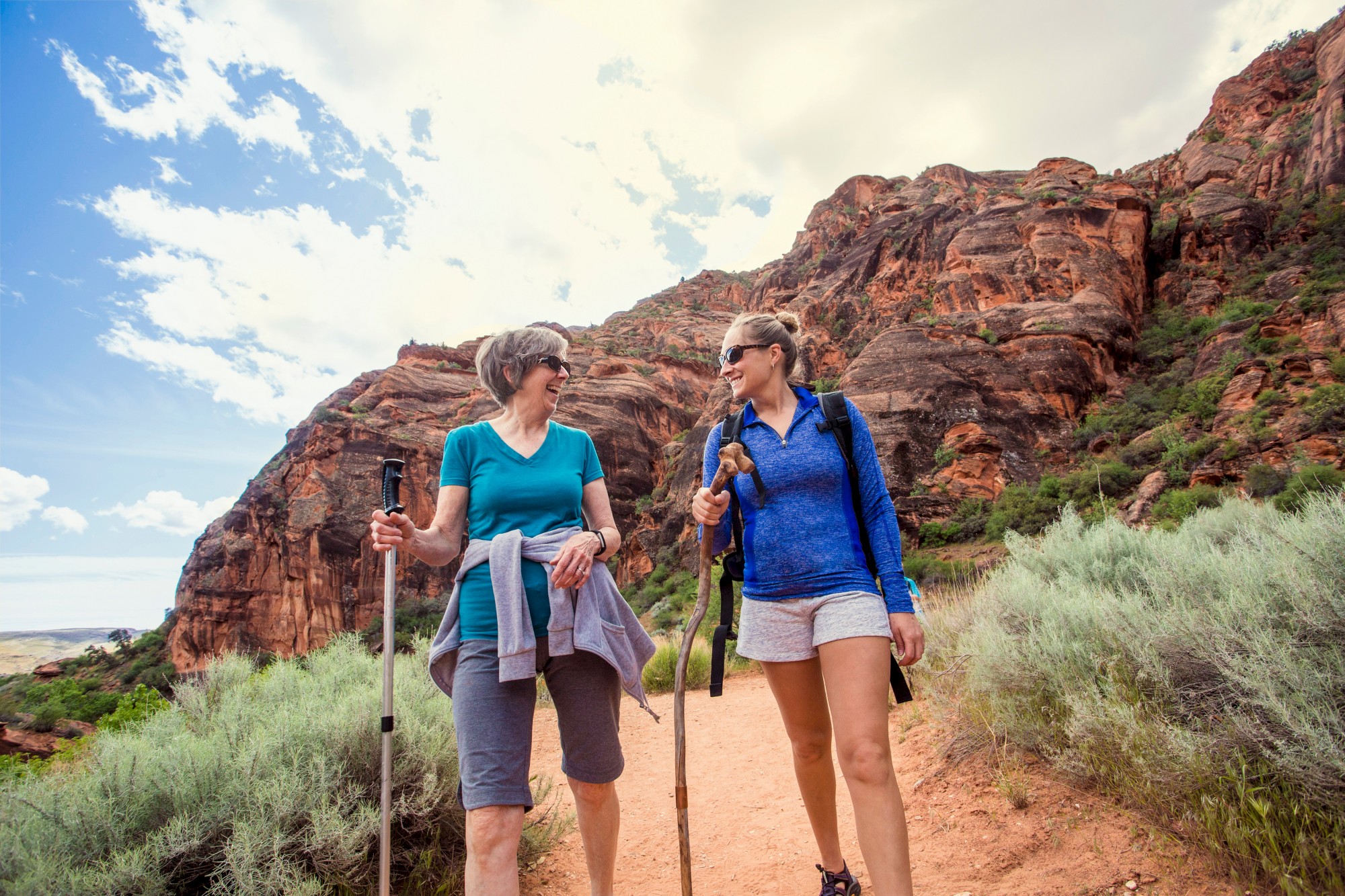 Two women with hiking poles walking on a desert trail, with rocky cliffs in the background and green vegetation around them.
