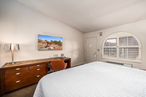 A tidy room with a bed, wooden desk, chair, wall art, and a lamp. It also features a door and arched window with shutters.