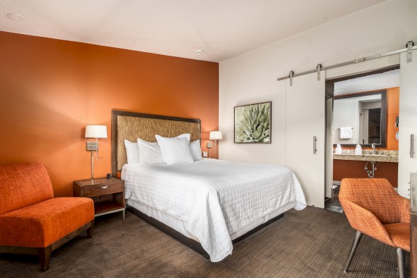 A hotel room with a bed, orange accent wall, chairs, nightstand, and a view into the bathroom with a sliding door and sink area.