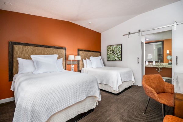 The image shows a hotel room with two beds, an orange wall, a desk with an orange chair, and a sliding door leading to a bathroom.