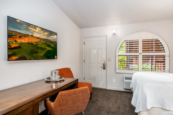 A hotel room features a TV, seating, and a desk with a water carafe. A window with wooden shutters allows natural light inside.