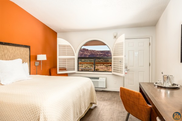 A hotel room with a bed, desk, and chair, featuring a window with shutters open to a scenic mountain view, and a bold orange wall.