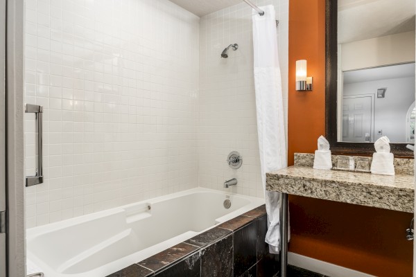 A bathroom featuring a white bathtub with a shower curtain, a granite countertop with towels, and a wall-mounted light fixture near a mirror.