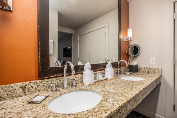 A bathroom with a granite countertop features dual sinks, a large mirror, wall sconces, toiletries, and folded towels, creating a neat appearance.