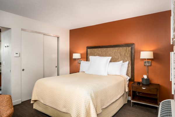 The image shows a tidy bedroom with an orange accent wall, a bed, side tables, wall lamps, and a wicker chair by a closet with sliding doors.