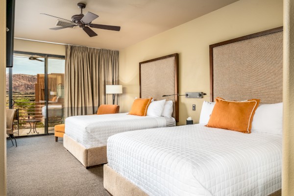 The image shows a hotel room with two beds, orange pillows, wall decor, and a balcony with a view of a mountainous landscape outside.
