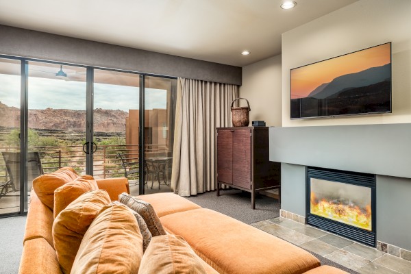 A cozy living room with an orange sofa, fireplace, wall-mounted TV, glass doors, and outdoor view of mountains on the patio.