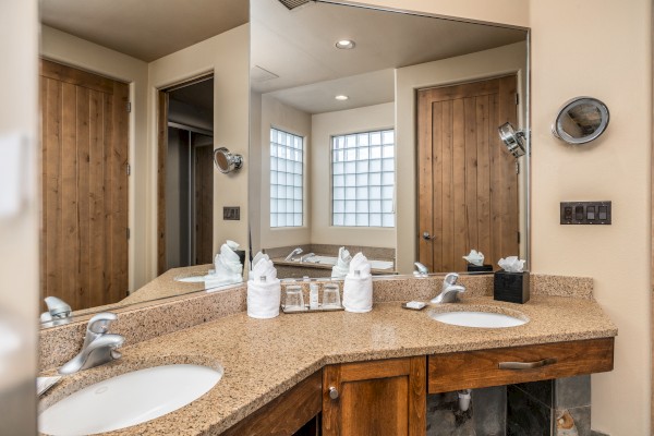 The image shows a bathroom with a double sink, granite countertops, mirrors, wooden doors, and folded towels on the counter.