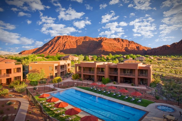 The image shows a resort with a pool, surrounded by desert landscape and red rock cliffs under a partly cloudy sky.
