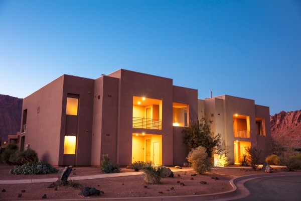 Modern desert home at dusk, warm lights on, surrounded by plants and rocks, with a clear sky and mountains in the background.