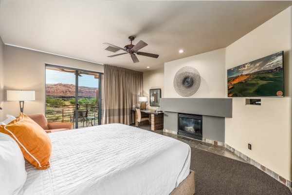 A hotel room features a bed, TV, fireplace, and a desk with a chair. There's a sliding door leading to a view of a desert landscape.