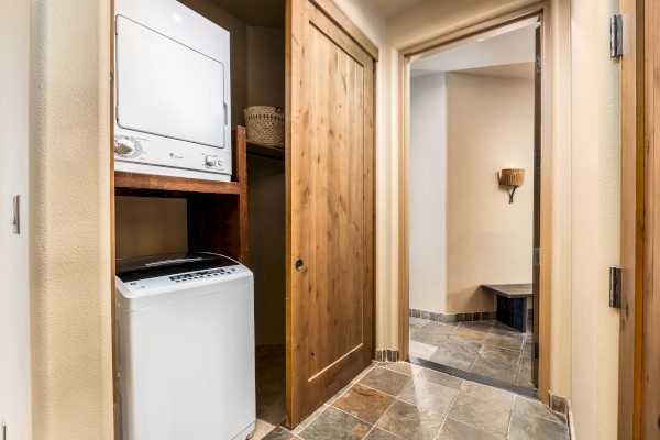 A hallway with a stacked washer and dryer in a wooden closet beside an open door, leading to a stone-tiled room with a bench.