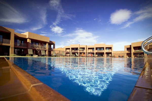 A modern outdoor swimming pool with reflections, surrounded by buildings under a clear blue sky with some clouds.