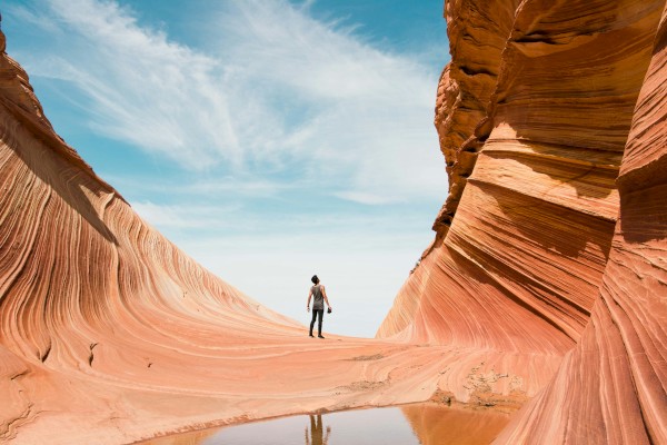 A person stands between colorful, wavy rock formations under a blue sky with clouds, near a small water pool.