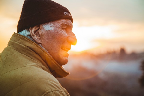 An elderly man wearing a beanie and jacket smiles while looking at a scenic sunrise or sunset, with a soft, blurred landscape in the background.