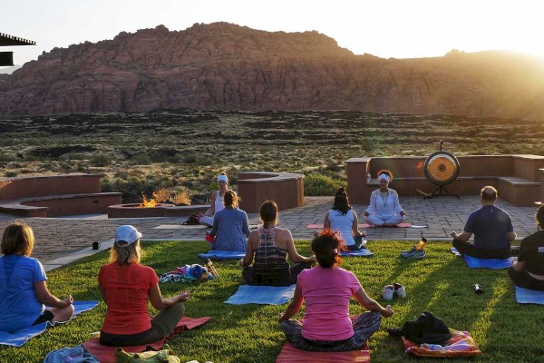 A group of people are practicing yoga outdoors on mats, facing an instructor, with a scenic desert landscape and setting sun in the background.