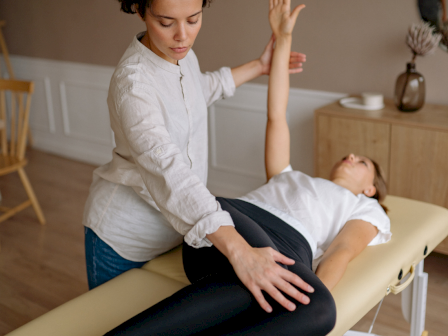 A person is assisting another with a stretch on a massage table in a calm room with gentle lighting.