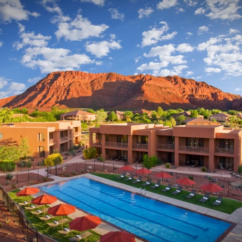 The image shows a resort with a swimming pool surrounded by modern buildings and scenic red rock formations under a blue sky.