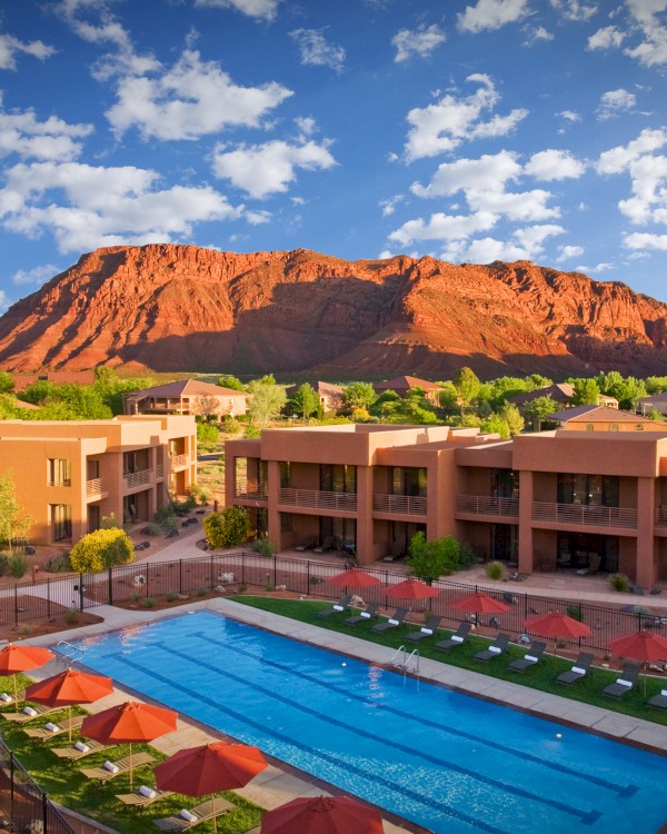 The image shows a resort with a swimming pool surrounded by modern buildings and scenic red rock formations under a blue sky.