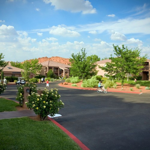 A peaceful suburban street with greenery, a child on a bike, two people walking, and buildings in the background under a blue sky with clouds.