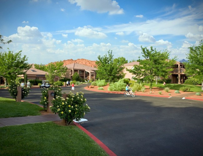 A peaceful suburban street with greenery, a child on a bike, two people walking, and buildings in the background under a blue sky with clouds.