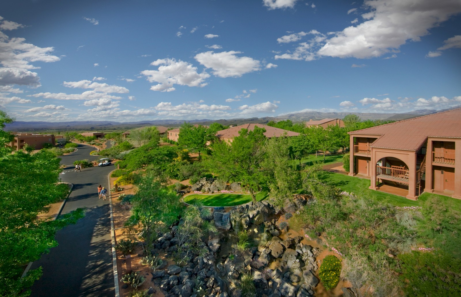 The image shows a peaceful residential area with houses, trees, a waterfall feature, and people walking on the street on a sunny day.