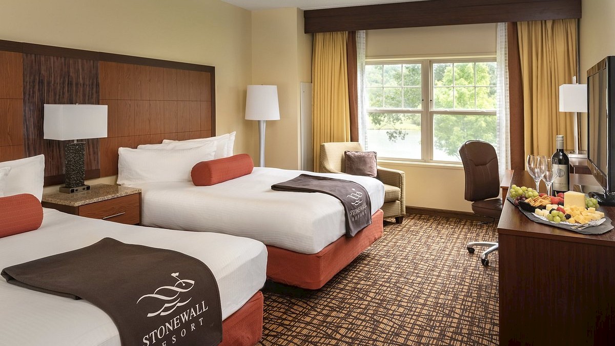 A hotel room with two double beds, a window, a desk with wine and fruit, and 