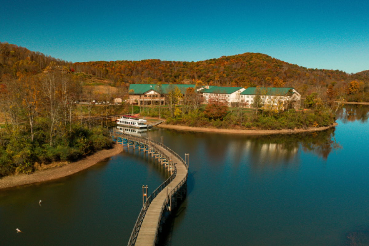 A scenic aerial view shows a peaceful lake with a long, winding wooden walkway leading to buildings surrounded by autumn trees and hills.