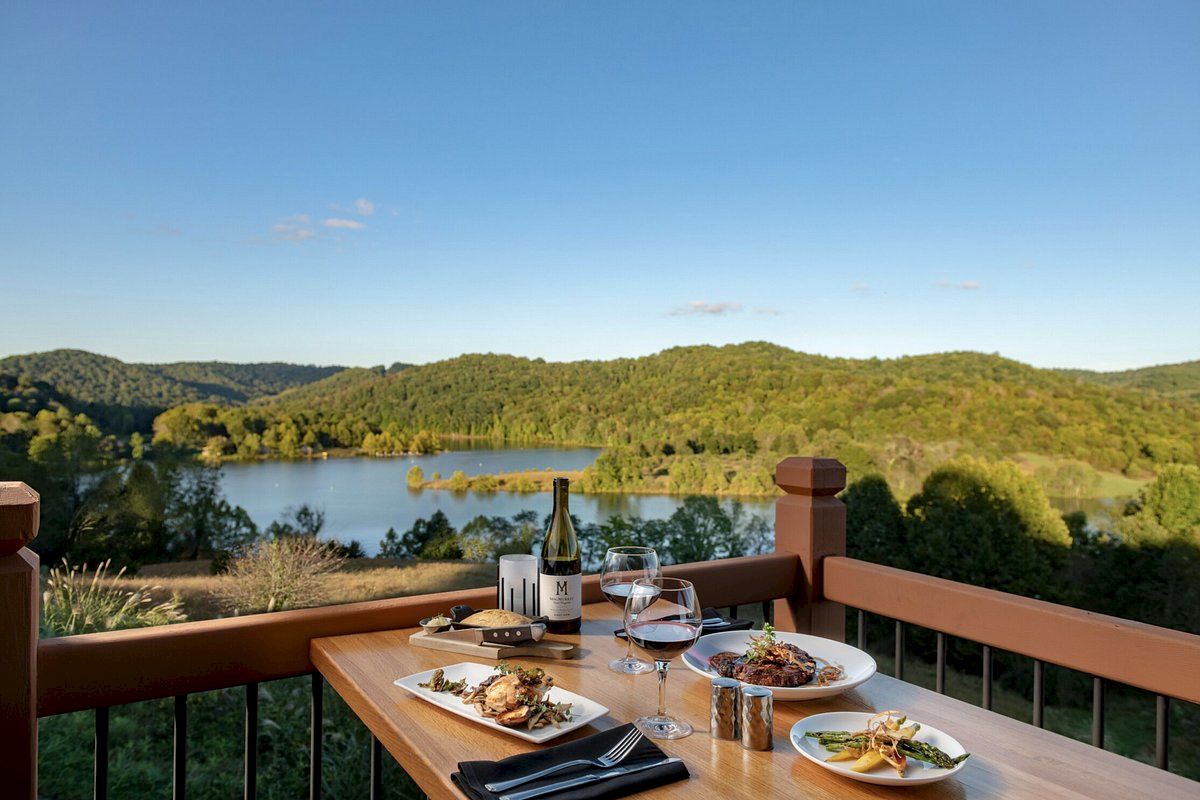 A table set with plates of food, wine glasses, and a bottle of wine is on a balcony overlooking a scenic, green landscape with a lake.