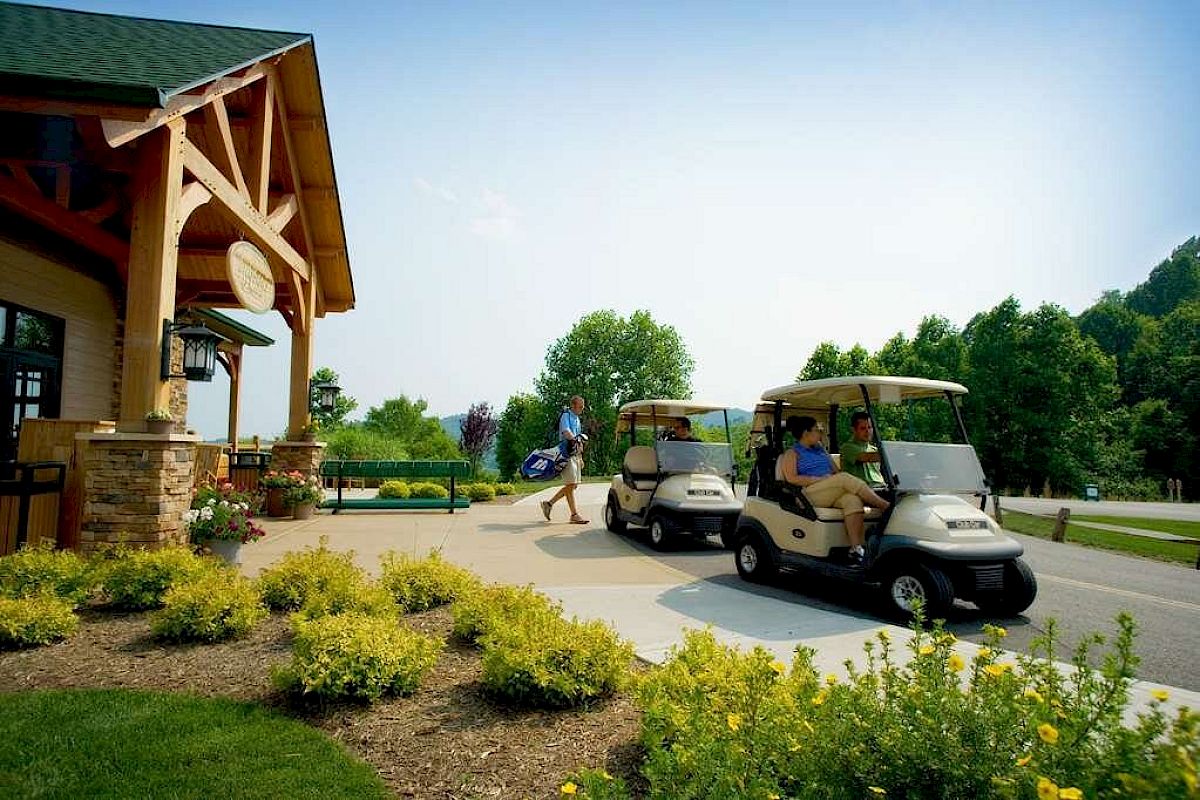 A person is walking towards a building, while two golf carts with passengers are on a path nearby, surrounded by greenery and flowers.