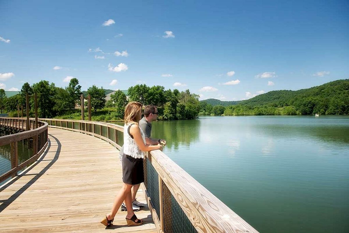 Two people stand on a wooden boardwalk over a lake, with hills and trees in the background, enjoying the scenic view under a clear blue sky.