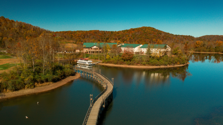 The image depicts a serene lakeside scene with a curving bridge connecting to a building complex surrounded by autumn-colored trees and hills, under a clear sky.