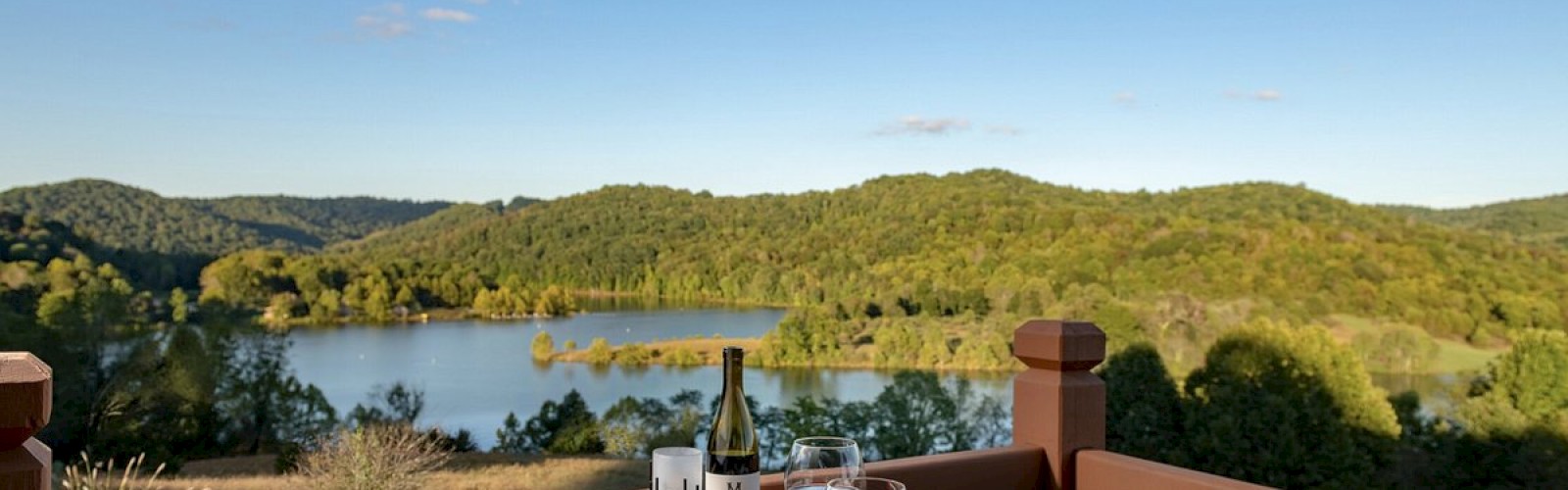 A dining table on a balcony with plates of food, wine, and glasses, overlooking a scenic lake and forested hills under a clear blue sky.